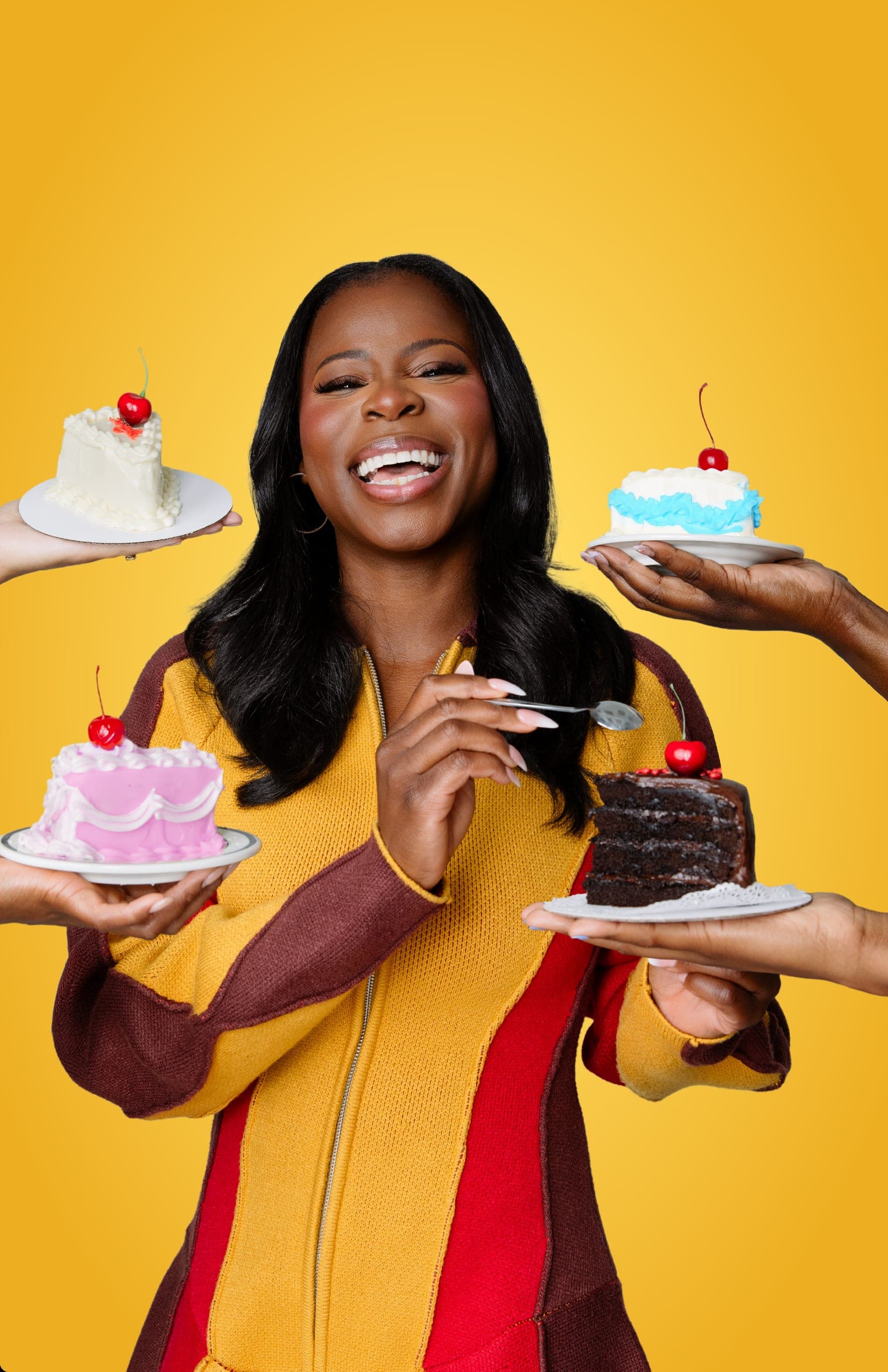 Manouchka Charles laughing, a spoon raised in one hand, in a mustard, maroon and red colour-blocked knit, with four outstretched hands around her offering small cherry-topped cakes — white, blue, pink, and a slice of chocolate — against a golden yellow backdrop.